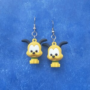 Pode incluir: Um par de brincos pendentes com pingentes de cão de desenho animado amarelo, com orelhas pretas, focinhos brancos e coleiras verdes. Os pingentes estão presos a ganchos de metal prateado por pequenos anéis, criando um acessório colorido.