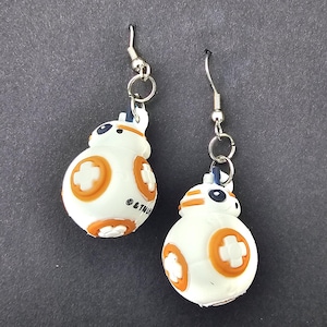 May include: A pair of drop earrings featuring spherical white and orange droid charms with silver-toned metal hooks. Each charm displays a circular orange pattern with a white cross in the centre and small black details. The text & TM is visible.