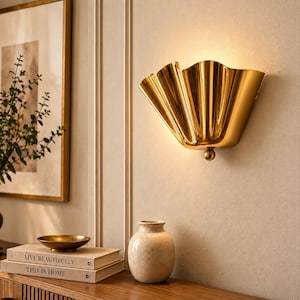 Może przedstawiać: Złoty kinkiet o plisowanym, wachlarzowatym kształcie z małym kulistym akcentem u dołu. Oprawa emituje ciepły, miękki blask z górnego otworu, rzucając światło w górę na powierzchnię ściany, tworząc jasne i odblaskowe wykończenie.