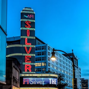 May include: A movie theater marquee with the words "AFI Silver Theatre" in red and blue neon lights. The theater is located in a city with tall buildings in the background.