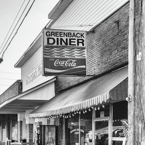 Puede incluir: Una foto en blanco y negro de un restaurante con un letrero que dice "Greenback Diner" y un letrero de Coca-Cola. El restaurante tiene un toldo a rayas y un poste de madera en frente.