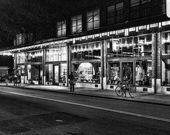 May include: A black and white photo of a storefront at night. The store has large windows and is lit up. There is a person standing in front of the store and a bicycle parked in front of the store.