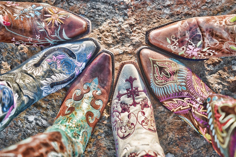 Op de afbeelding: Vijf paar kleurrijke, geborduurde cowboylaarzen met ingewikkelde bloem- en geometrische patronen. De laarzen zijn in een cirkel op een rotsachtig oppervlak gerangschikt.