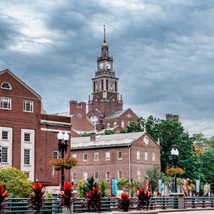 May include: A brick building with a clock tower and a cloudy sky. The building has a clock tower with a clock face and a spire. The building is surrounded by trees and bushes. There are several potted plants in front of the building.