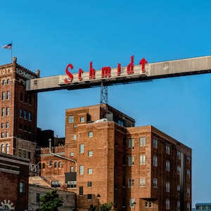 May include: A red neon sign with the word "Schmidt" on a metal structure over a brick building. The building has a large, white silo to the right.
