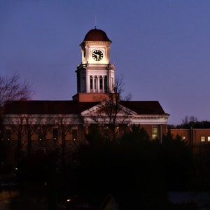 May include: A brick building with a clock tower at dusk. The clock tower has a white dome and a clock face with Roman numerals. The building has a red roof and white trim.