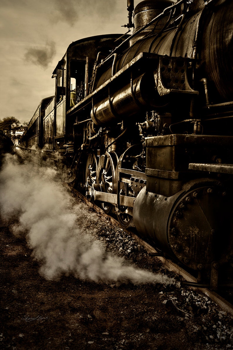 May include: A vintage steam locomotive train engine with black wheels and a large smokestack, emitting white smoke, traveling on a dirt track.