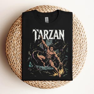 May include: A black short-sleeved t-shirt featuring a graphic print of the character Tarzan in a dynamic pose. The word TARZAN is printed in large white distressed lettering above the character, who is surrounded by jungle foliage and vines.