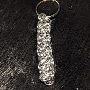 May include: A silver-colored metal keychain featuring a woven chainmail pattern constructed from small, interlocking circular rings. The rectangular-shaped chainmail section is attached to a standard circular metal split ring at the top.