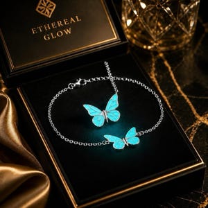 May include: A silver-toned chain bracelet featuring two glowing cyan butterfly charms. The jewelry is displayed inside a black box with the text ETHEREAL GLOW printed in gold on the lid. The butterflies have intricate wing details.
