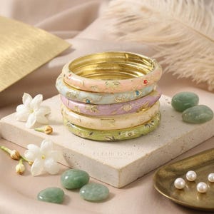 May include: A stack of five round bangles in pastel shades of pink, light blue, lavender, cream, and sage green. Each bangle features a gold-tone floral pattern and a smooth, glossy finish. The bracelets are stacked vertically on a stone block.