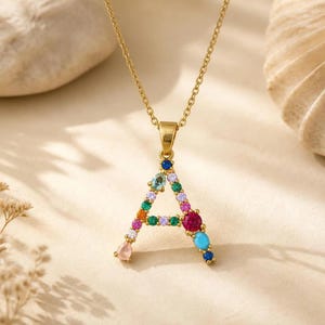 May include: A gold-tone necklace featuring a pendant shaped like the letter A. The pendant is embellished with an assortment of multi-colored, round, and pear-shaped gemstones, including shades of blue, green, pink, orange, and clear white stones.