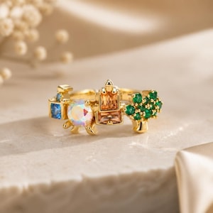 Puede incluir: Un anillo de tono dorado que presenta una serie de elementos decorativos, incluyendo una pequeña piedra cuadrada azul, una piedra redonda iridiscente, una piedra rectangular naranja central y un grupo de pequeñas piedras verdes en forma de árbol.
