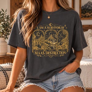 May include: A charcoal gray short-sleeve t-shirt featuring a gold graphic print. The design includes three arched panels depicting fantasy landscapes with text that reads I AM A SURVIVOR OF MAAS DESTRUCTION, CRESCENT CITY, TERRASEN, and VELARIS.