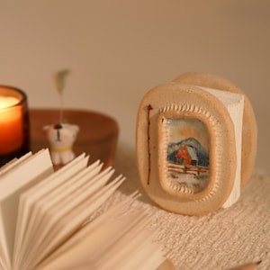 Può includere: Un piccolo oggetto in ceramica beige, di forma rotonda, con un bordo testurizzato e un paesaggio dipinto a mano raffigurante una casa rossa, montagne e una recinzione. L'oggetto è posizionato in verticale accanto a un libro aperto.