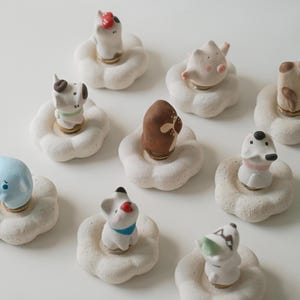 Puede incluir: Una colección de nueve pequeñas figuras de cerámica, cada una con un animal único sobre una base blanca texturizada en forma de flor. Los animales incluyen varios perros, un cerdo y un elefante azul, todos con detalles pintados a mano.
