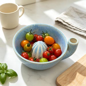 May include: A light blue ceramic citrus juicer with a speckled glaze finish, featuring a central reamer and a side pouring spout. The bowl is filled with a variety of fresh, multi-coloured cherry tomatoes in shades of red, orange, and green.