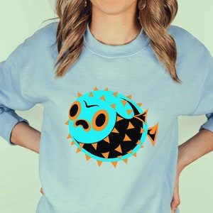 May include: A light blue sweatshirt with a graphic of a blue and black pufferfish with orange spikes. The pufferfish has a cartoon-like face with large eyes and a frown.