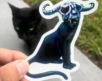 Könnte beinhalten: Ein schwarzer Katzenaufkleber mit einem Schädel und Hörnern auf dem Kopf. Die Katze sitzt mit ihrem Schwanz um den Körper gewickelt. Der Aufkleber hat einen weißen Hintergrund.
