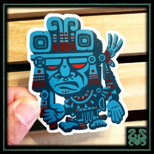 May include: A die-cut vinyl sticker featuring a stylized illustration of a Mesoamerican-inspired deity figure. The design uses a bold color palette of bright cyan blue and deep red accents, depicting a figure with an ornate headdress.