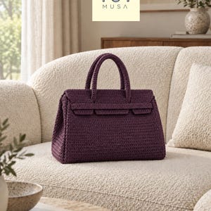 Peut inclure: Un sac à main violet foncé réalisé avec un point de crochet dense, doté de deux anses supérieures et d'une sangle horizontale sur le devant. Le sac présente une silhouette rectangulaire structurée avec un rabat et le logo MUSA inscrit.