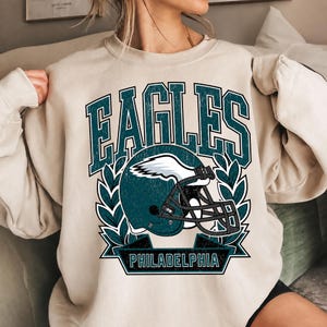 May include: A beige crewneck sweatshirt featuring a graphic print with the word EAGLES in large teal letters above a teal football helmet with a white wing design. Below the helmet is a teal banner with the word PHILADELPHIA in teal letters.
