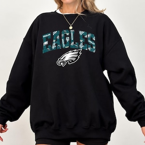 Peut inclure: Un sweat-shirt noir à manches longues et col rond arborant le mot EAGLES dans une grande police arquée de couleur sarcelle avec un motif répétitif. Sous le texte se trouve un logo représentant une tête d'aigle blanche, noire et sarcelle.