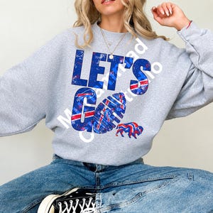 Pode incluir: Uma camisola cinzenta clara de gola redonda com o texto LET'S GO num padrão desgastado azul, vermelho e branco. O design inclui um gráfico de uma bola de futebol americano e uma silhueta de bisão, ambos com as mesmas cores.
