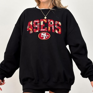 Könnte beinhalten: Ein schwarzes Langarm-Sweatshirt mit dem Schriftzug 49ERS in großen, roten Buchstaben mit Used-Optik. Die Buchstaben enthalten kleine weiß-rote Grafiken zum Thema Football. Darunter befindet sich ein rot-gold-weißes 49ers-Teamlogo.