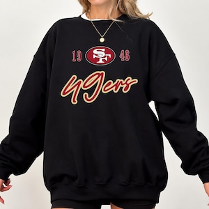 Könnte beinhalten: Ein schwarzes Langarm-Sweatshirt mit Rundhalsausschnitt und dem Logo der San Francisco 49ers in Rot, Gold und Weiß. Die Grafik zeigt den Text 1946 und das Wort 49ers in einer kursiven Schriftart mit goldener Umrandung und roter Füllung.