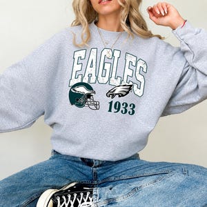 Puede incluir: Una sudadera gris claro de cuello redondo con la palabra EAGLES en letras blancas grandes y desgastadas. Debajo del texto hay un casco de fútbol americano verde oscuro con un gráfico de ala blanca, un logo de águila y el número 1933 en verde.