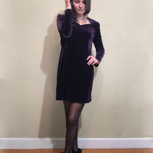 May include: A woman wearing a purple velvet dress with long sleeves and a square neckline. She is wearing black tights and black ankle boots.