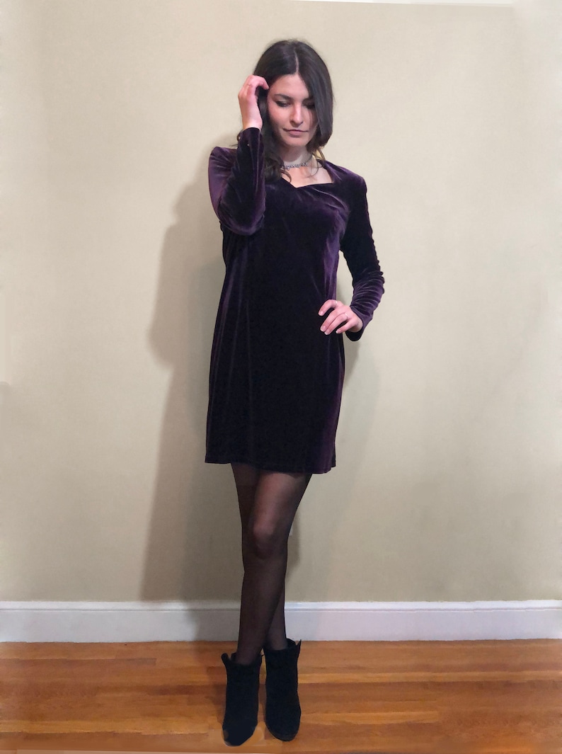 May include: A woman wearing a purple velvet dress with long sleeves and a square neckline. She is wearing black tights and black ankle boots.
