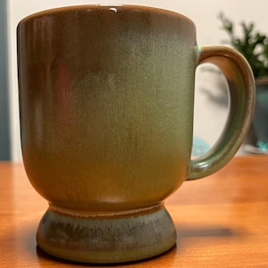 May include: A ceramic mug featuring a smooth, glazed finish in shades of olive green and brown. The mug has a cylindrical body, a curved handle on the right side, and a distinct pedestal base that flares slightly at the bottom edge.