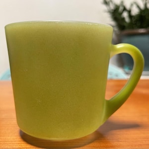 May include: A translucent light green glass mug with a smooth, matte-like finish and a curved handle. The mug features a cylindrical body with a slightly tapered base and a consistent, solid lime-green color throughout the entire glass structure.