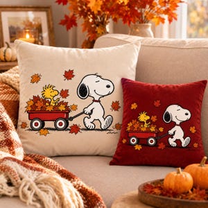 May include: Two square throw pillows featuring Snoopy pulling a red wagon filled with autumn leaves and Woodstock. One pillow is cream-colored, and the other is deep red. Both display the same cartoon illustration of the characters.