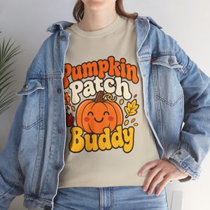 Puede incluir: Una camiseta beige con un gráfico de una calabaza naranja sonriente y el texto Pumpkin Patch Buddy en letras retro naranjas y amarillas, rodeado de pequeñas hojas de otoño, usado debajo de una chaqueta de mezclilla azul claro y jeans azules.