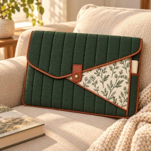 May include: A rectangular laptop sleeve featuring a dark green quilted fabric exterior with vertical stitching. It has a brown leather-like trim, a central snap button closure, and a triangular side panel with a cream background and black floral print.