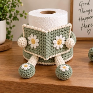 Puede incluir: Una funda de ganchillo para papel higiénico en color verde salvia, con un panel frontal en forma de libro y aplicaciones de margaritas blancas. Incluye dos piernas colgantes con pies decorados con margaritas y dos pequeños brazos redondos.
