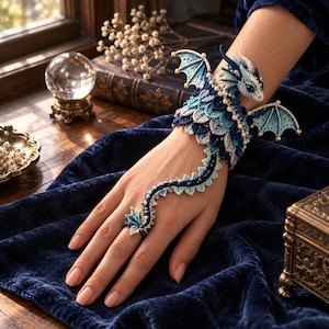 Puede incluir: Una pulsera de dragón tejida a ganchillo con escamas superpuestas en azul y blanco, alas intrincadas y una cola larga que se extiende hasta un anillo. El diseño está adornado con pequeñas cuentas blancas a lo largo del cuerpo y las alas.