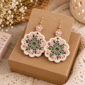 May include: A pair of round crochet earrings featuring a sage green center with small pink flowers and white pearl accents. The earrings have a scalloped cream-colored border and are attached to gold-toned metal hooks with a small pink flower.