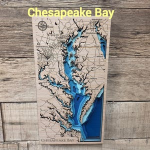May include: A rectangular wooden wall map of the Chesapeake Bay featuring laser-cut topographical layers in varying shades of blue to represent water depth, with black engraved land details, city names, and a compass rose on a light wood surface.