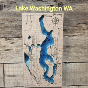 May include: A rectangular wooden bathymetric map of Lake Washington, WA, featuring layered blue water depths carved into light-colored wood. The map displays surrounding land topography, road networks, a compass rose, and the text Lake Washington WA.