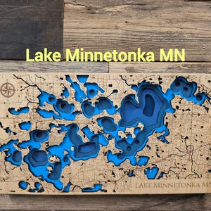 Puede incluir: Un mapa batimétrico rectangular de madera del lago Minnetonka MN con recortes de madera clara en capas, detalles de líneas negras intrincadas y profundidades de agua azul intenso. El texto Lake Minnetonka MN está grabado en la esquina.