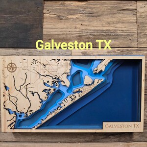 May include: A rectangular wooden wall art piece featuring a layered bathymetric map of Galveston TX. The light wood top layer displays detailed land topography and a compass rose, while the recessed blue layers represent water depths.