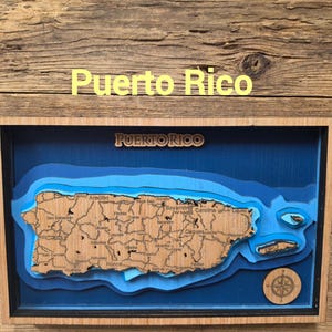 Op de afbeelding: Een rechthoekig houten wanddecoratie met een gelaagde 3D-kaart van Puerto Rico. De kaart toont natuurlijke houttinten met zwarte tekst voor plaatsnamen, geplaatst tegen diepblauwe waterlagen. Een kompasroos en de tekst Puerto Rico zijn aanwezig.
