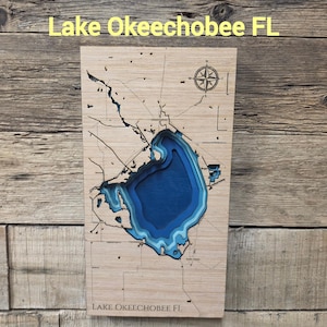 May include: A rectangular wooden wall art piece featuring a laser-cut bathymetric map of Lake Okeechobee FL. The map displays layered blue depths within the lake, surrounded by light wood grain, with a compass rose and the text Lake Okeechobee FL.