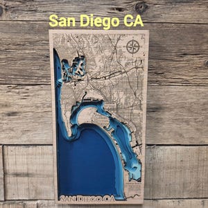 May include: A rectangular wooden wall art piece featuring a layered 3D bathymetric map of San Diego, CA. The map displays intricate land topography in light wood tones and deep blue water depths, with the text San Diego CA at the top and bottom.