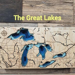 May include: A wooden bathymetric map of The Great Lakes featuring layered, laser-cut wood with blue-tinted water depths. The map includes a compass rose, labeled cities, and black engraved roads, with the text The Great Lakes printed in the corner.