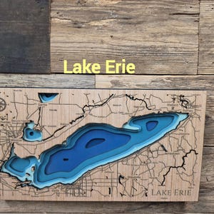 May include: A rectangular wooden wall map of Lake Erie featuring layered blue cutouts to represent water depth. The light-colored wood surface displays black engraved geographic details, roads, and city names, with the text Lake Erie printed.